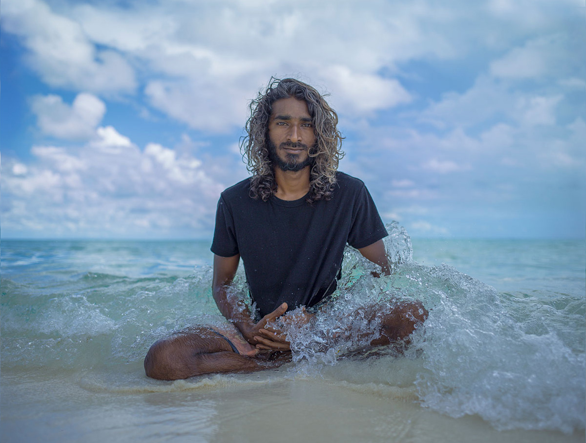 A photo of a man sitting waist deep in the ocean. He is wearing a black t shirt and has long hair.