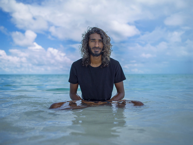 A photo of a man sitting waist deep in the ocean. He is wearing a black t shirt and has long hair.