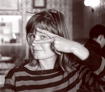 Young Pema Chodron looking at the camera. She has one eye peeking through a peace sign made with two of her fingers. She has bangs and shoulder-length hair and is wearing a striped long sleeve shirt.